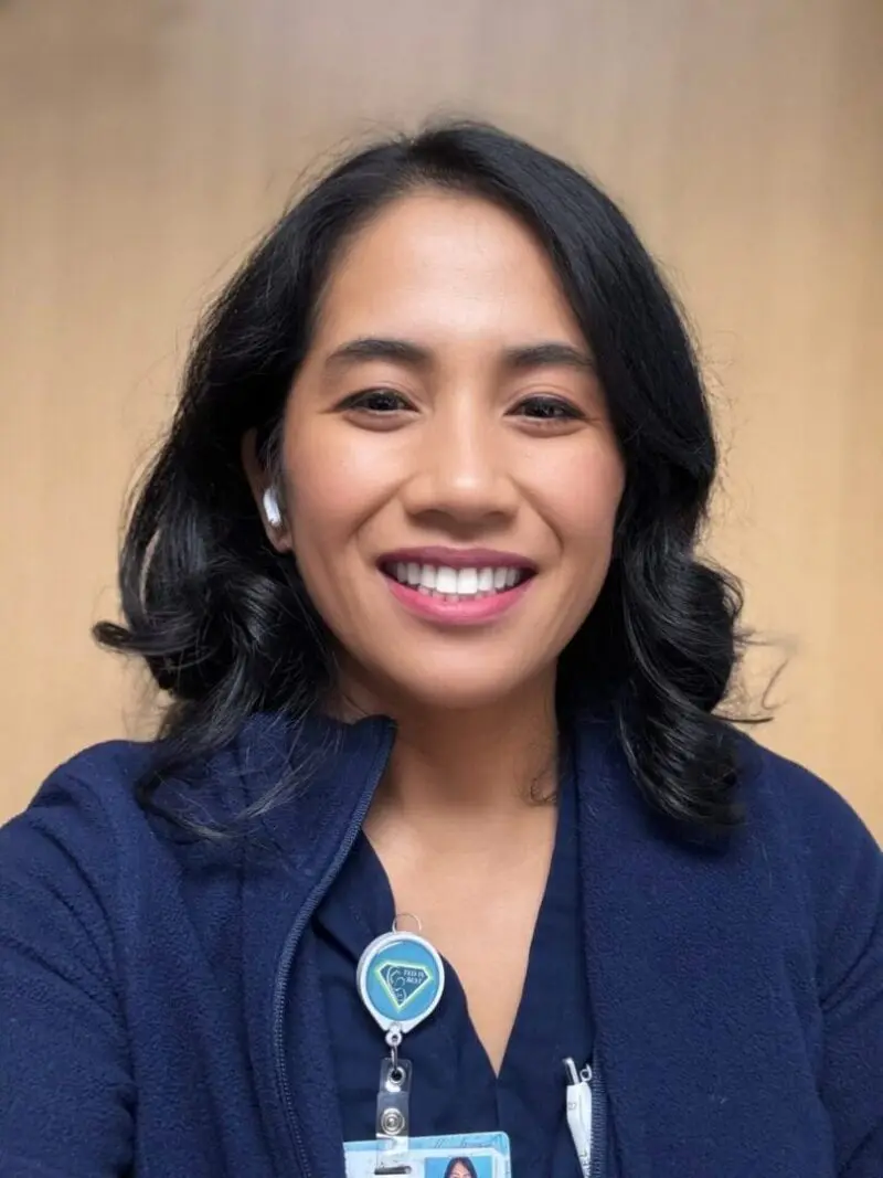 Dr. Christie del Castillo-Heygi smiling in front of a tan background wearing a navy blue top and wearing a fed is best badge reel